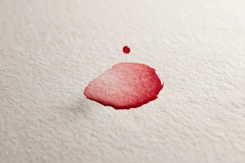 Nežna kapljica rjavkasto-rdeče barve na belem papirju ponazarja kako izgleda prva menstruacija